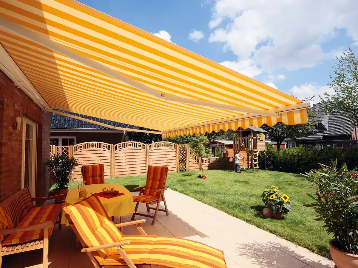 Eine sonnige Terrasse mit einer gestreiften Markise, die Schatten spendet, ausgestattet mit Holzstühlen und einem Tisch.